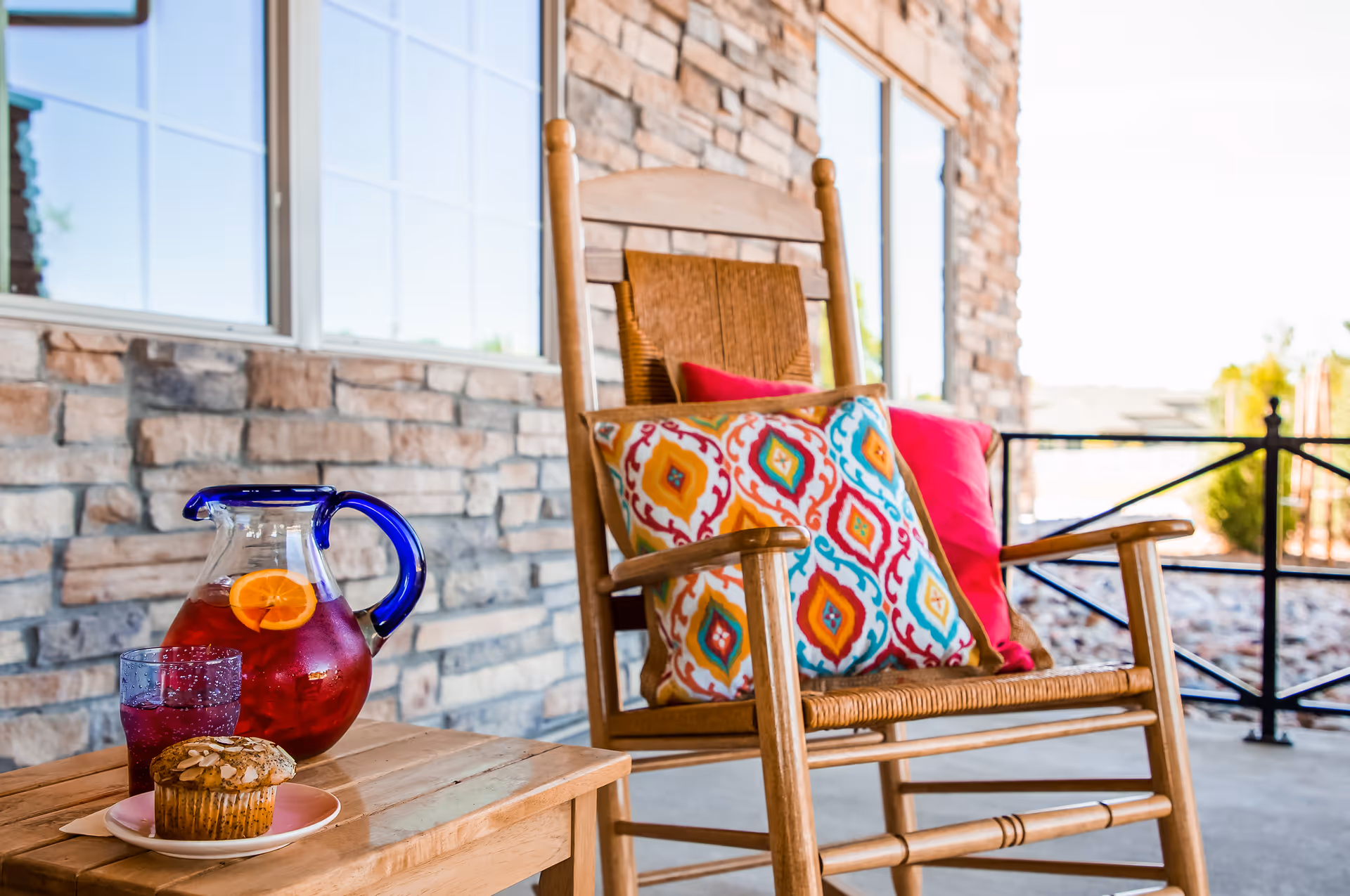 A wooden rocking chair with colorful patterned and solid red cushions on a porch with stone walls and large windows. A wooden table next to the chair holds a glass pitcher of red beverage with orange slices, a glass of the same drink, and a muffin on a white plate.