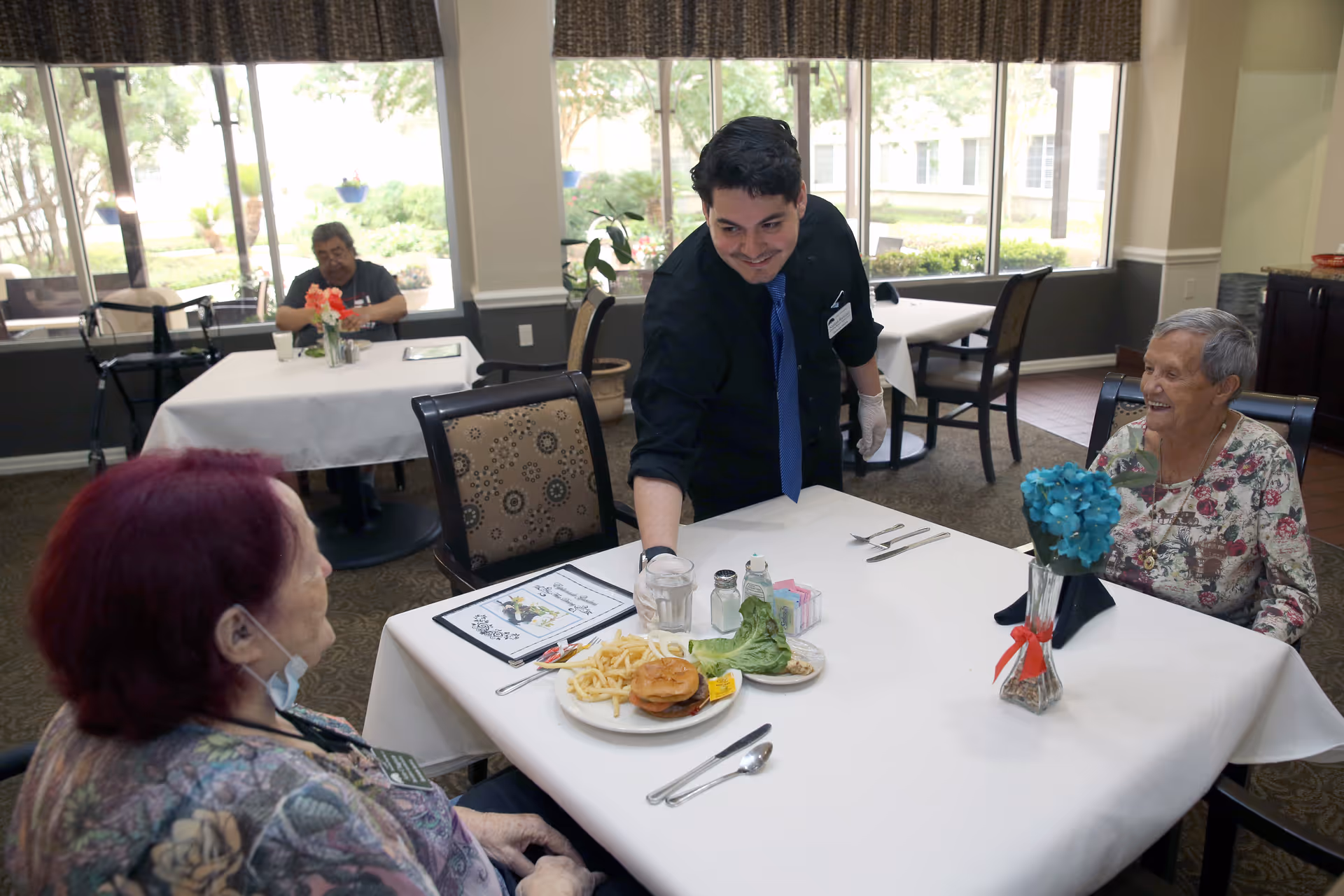 A staff member serves two elderly women seated at a table in a bright dining room with large windows.