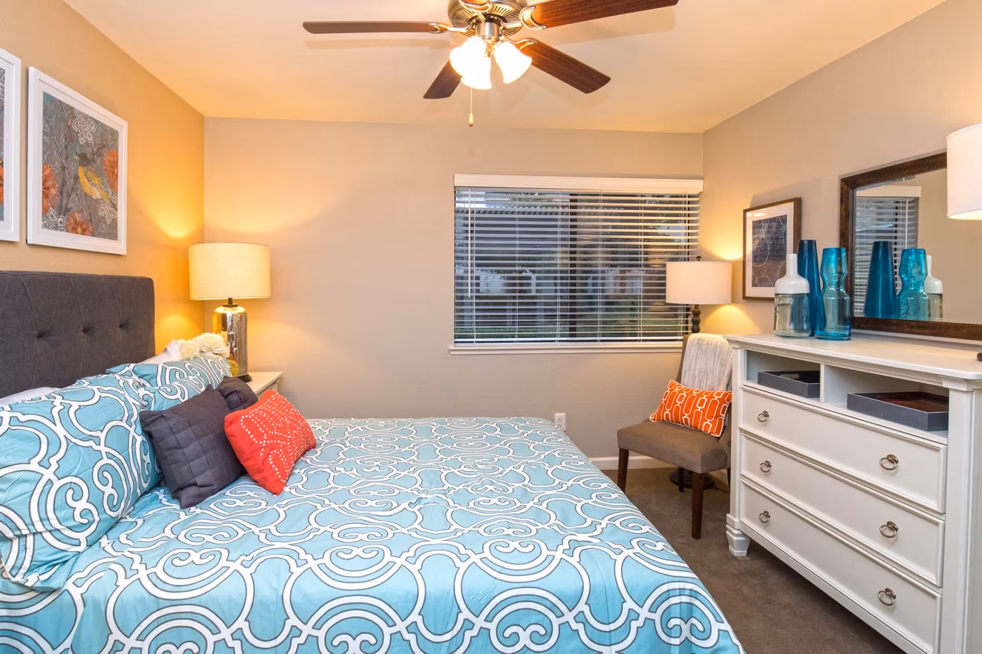 A cozy bedroom with a large bed covered in a blue and white patterned bedspread, multiple pillows including red and dark blue ones, a gray upholstered headboard, two bedside lamps, a window with blinds, a white dresser with decorative blue glass vases, a mirror above the dresser, a gray chair with an orange pillow, and framed artwork on the walls.