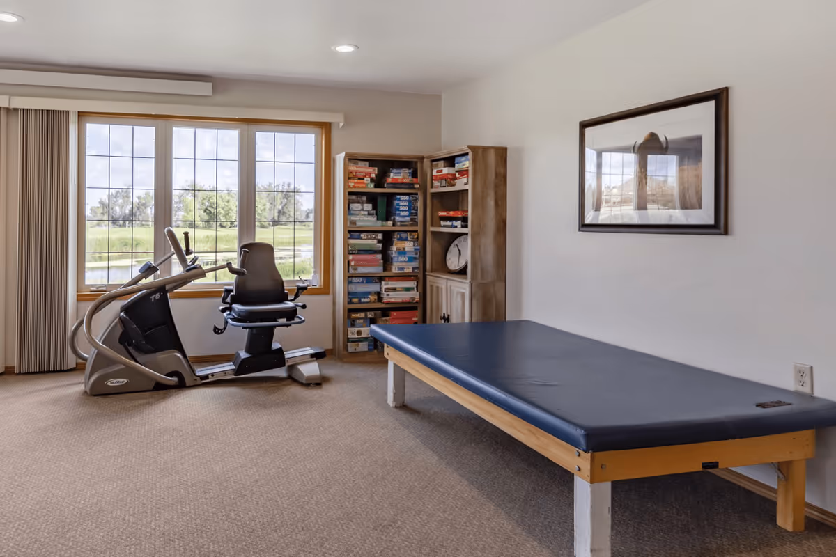 Interior activity room with a recumbent exercise machine, a padded therapy table, a bookshelf full of games, and large windows overlooking a grassy view.