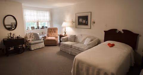 A cozy room with a single bed covered in a white blanket and a red heart-shaped pillow on top. Next to the bed is a floral-patterned sofa and a beige armchair. A wooden side table with a lamp is placed between the sofa and armchair. A window with white curtains lets in natural light, and a round mirror and framed artwork decorate the walls.