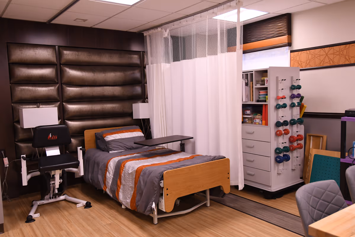 A rehabilitation room featuring a hospital bed with gray and orange bedding, a black padded chair with arm supports, a white privacy curtain, and a white storage cabinet holding colorful dumbbells and various supplies. The room has wood flooring and padded brown wall panels.