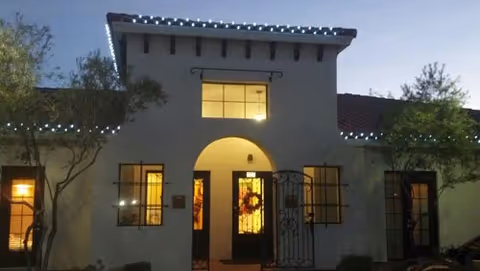 Front exterior of a Spanish-style building at dusk with a lit arched entrance, wreath on the door, and string lights along the roofline.