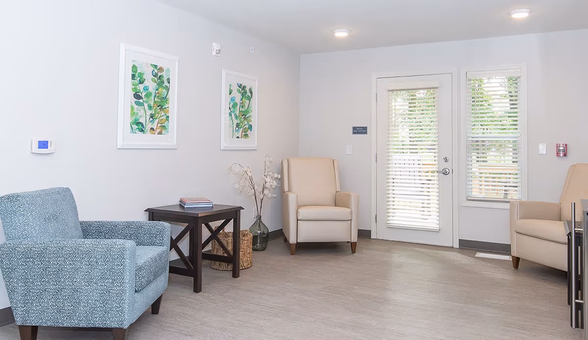 A bright and clean living room area with light gray walls and wood flooring. The room features two beige armchairs and one blue patterned armchair arranged around a dark wooden side table with books on it. Two framed botanical prints hang on the wall above the side table. A glass vase with decorative branches is placed on the floor next to the table. There is a door with white blinds and a window with matching blinds letting in natural light.
