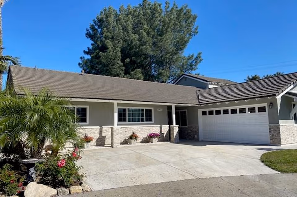 Single-story house with a gray roof, light gray exterior walls, and a two-car garage. There are several potted plants with flowers along the front porch and a small garden with green shrubs and pink flowers on the left side. Tall trees are visible in the background under a clear blue sky.