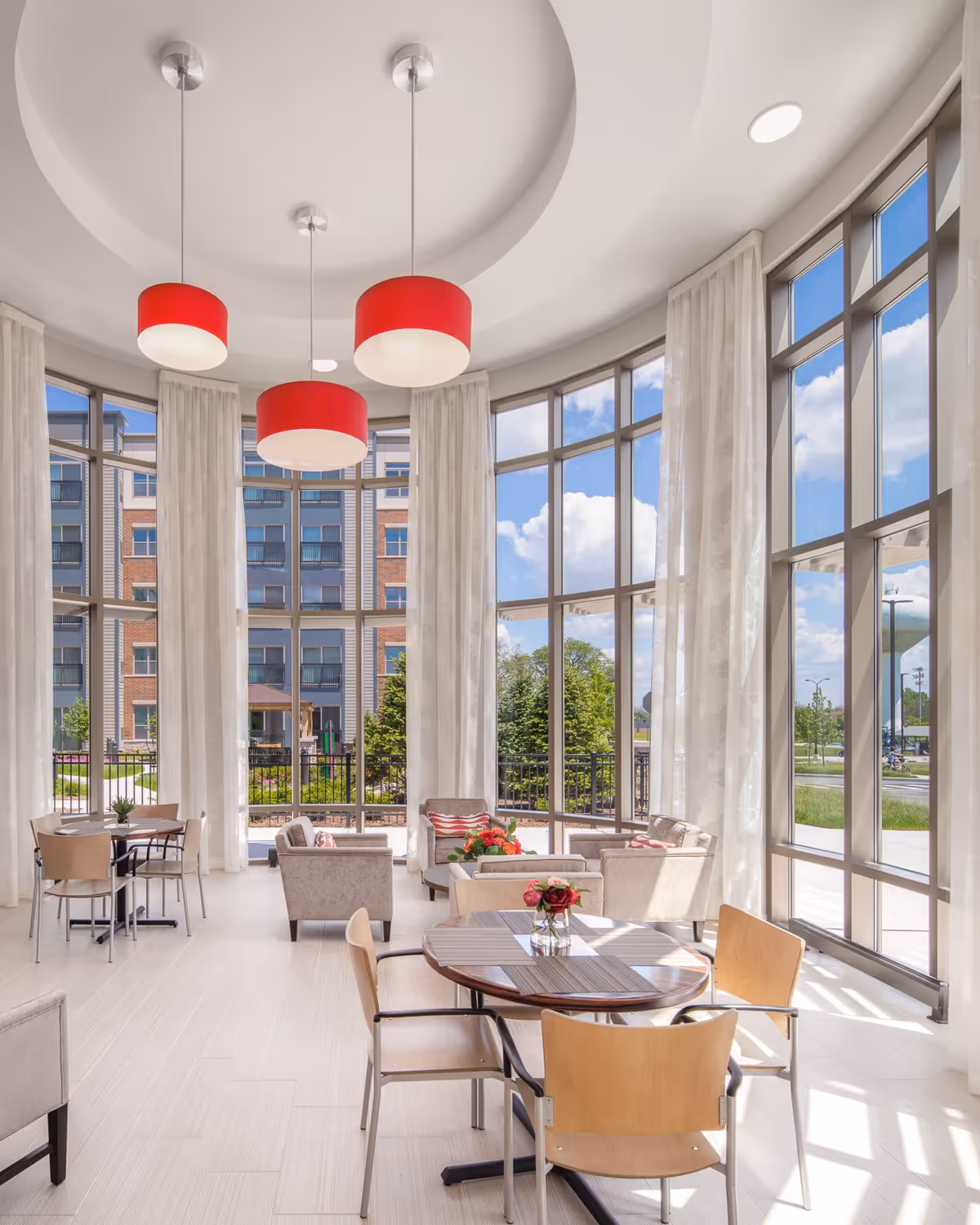 Bright and spacious common area with large floor-to-ceiling windows letting in natural light. The room features round tables with wooden chairs, comfortable armchairs with cushions, and red pendant lights hanging from a circular recessed ceiling. Outside the windows, a multi-story building and greenery are visible.