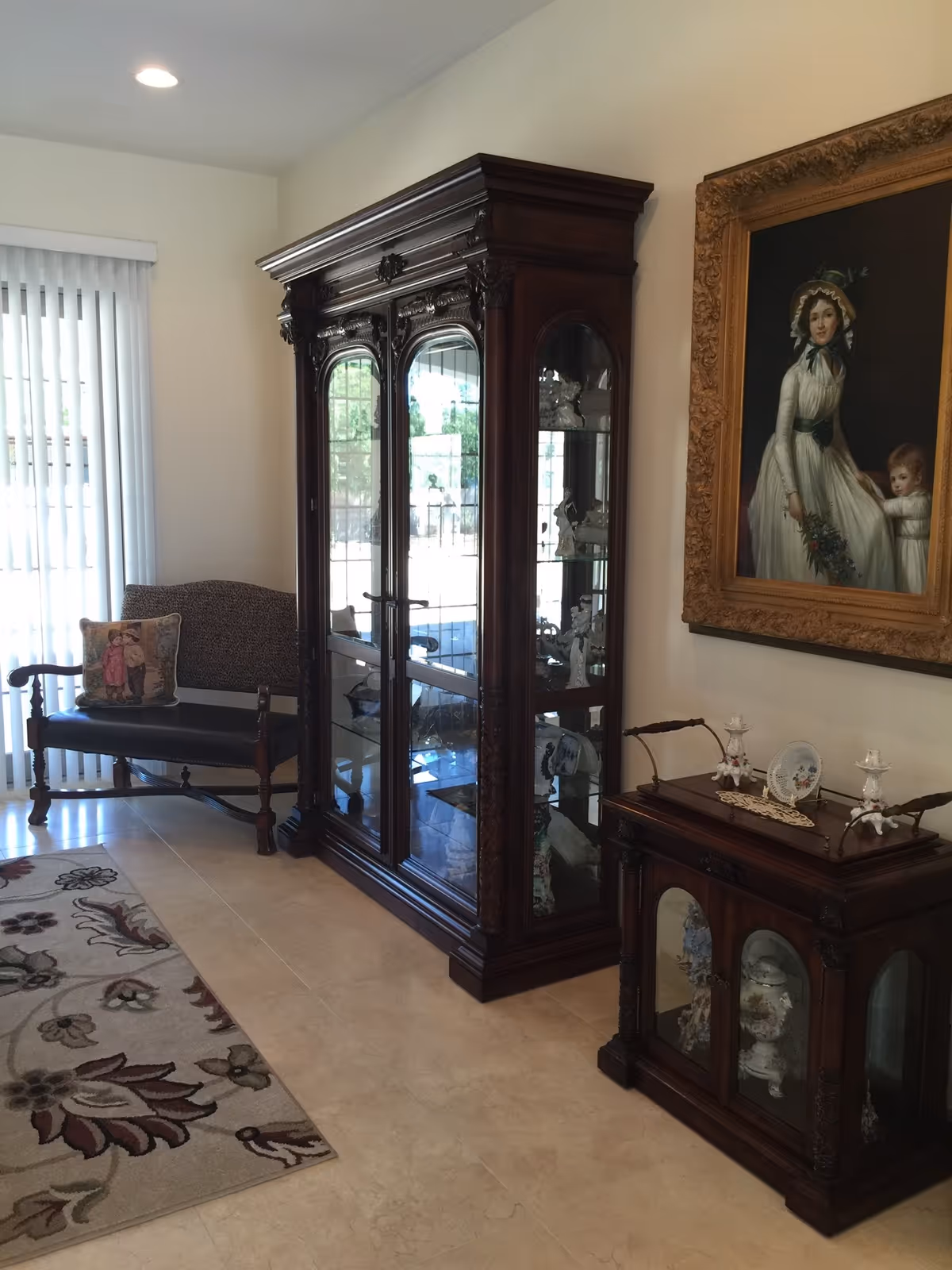 A living room area with a large wooden display cabinet filled with decorative items, a wooden bench with a cushion featuring a vintage image, a floral patterned rug on a tiled floor, and a framed portrait of a woman and child on the wall.