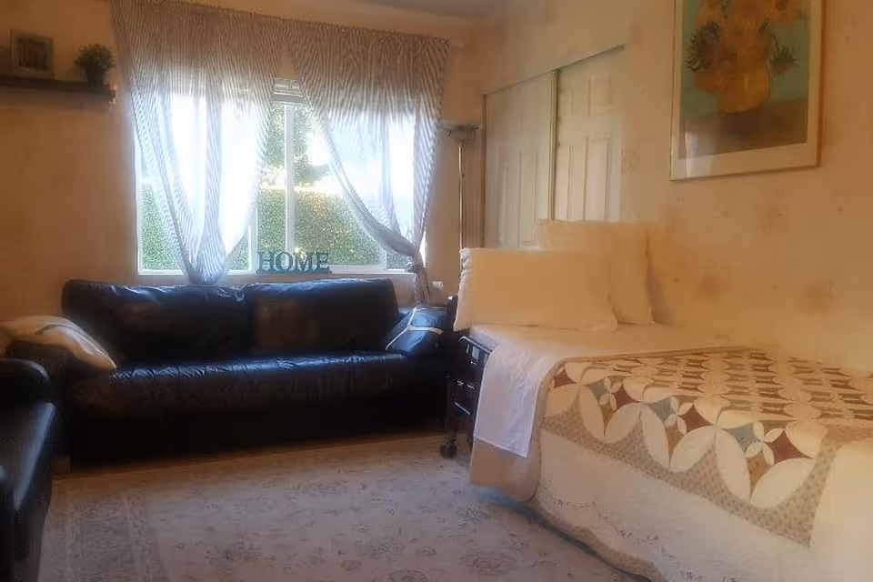A cozy room with a single bed covered with a patterned quilt and white pillows, a black leather sofa, a window with sheer curtains letting in natural light, a small side table with a lamp, and a framed painting on the wall.