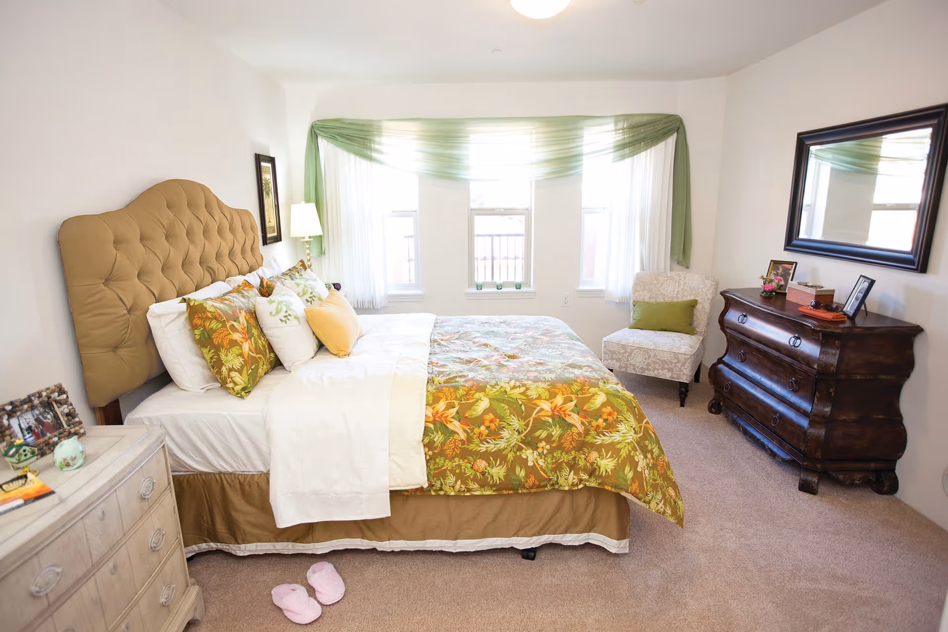 A cozy bedroom with a large bed featuring a tufted beige headboard and floral bedding in green, orange, and yellow tones. The bed is adorned with multiple pillows. There is a white nightstand with framed photos and decorative items on the left side of the bed. A pair of pink slippers is placed on the carpeted floor beside the bed. Three windows with sheer white curtains and green drapes allow natural light into the room. On the right side, there is a patterned armchair with a green cushion and a dark wooden dresser with a large mirror above it, decorated with framed photos and small items.