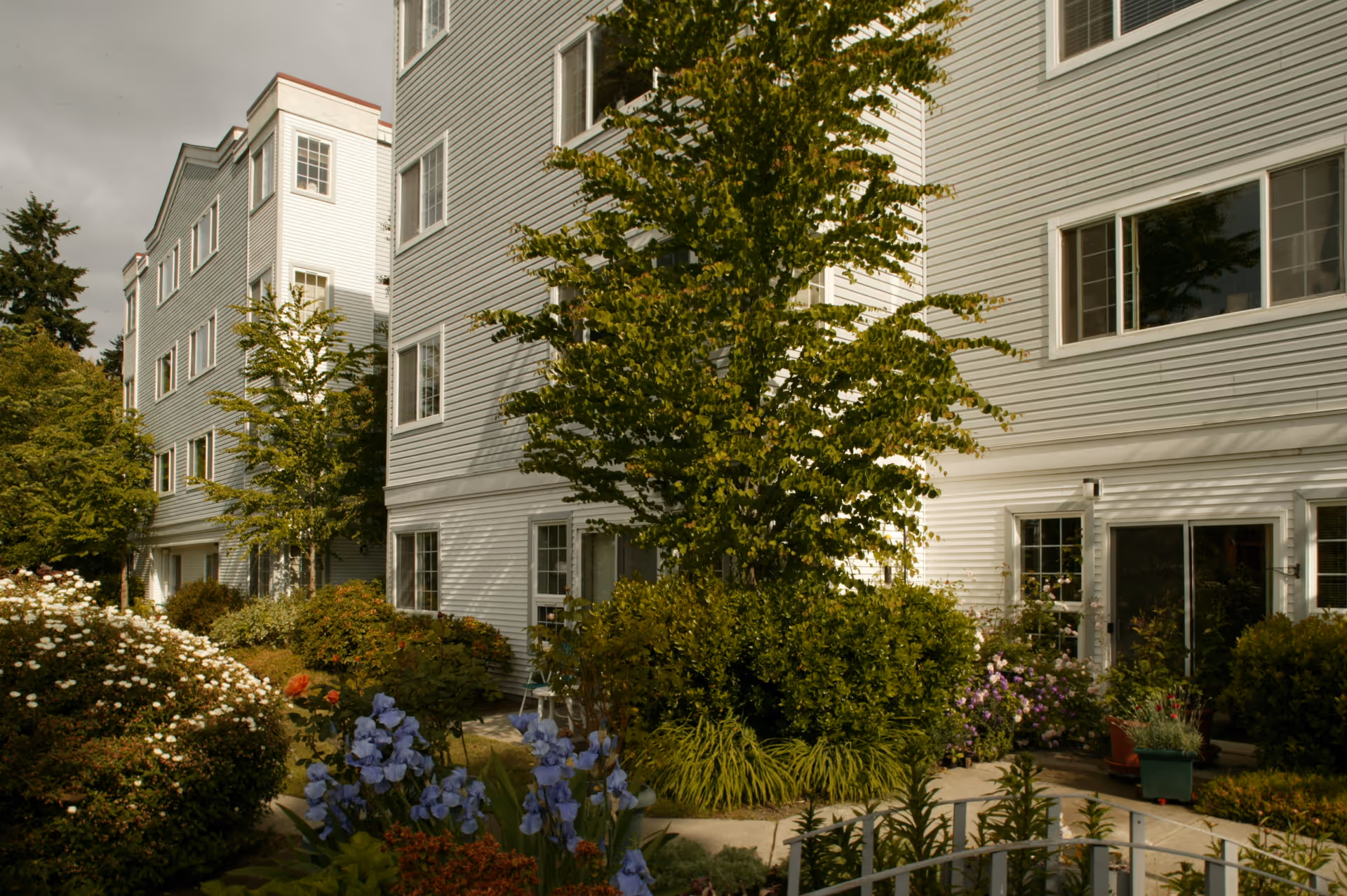 Exterior view of a multi-story senior living facility building with light gray siding, surrounded by lush greenery, flowering plants, and trees under a cloudy sky.