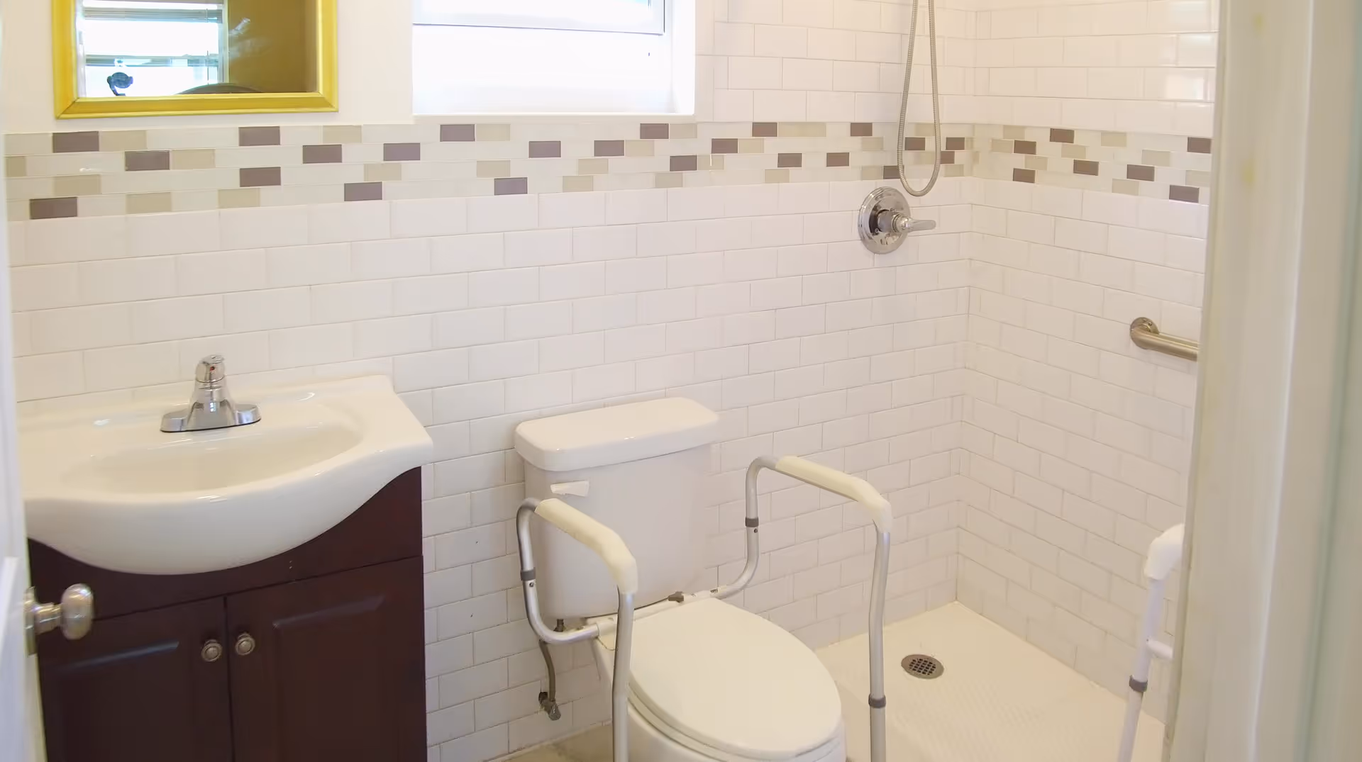 A bathroom with white tiled walls featuring a decorative border of beige and brown tiles. There is a white toilet with safety rails on both sides, a white sink with a dark brown cabinet below, a mirror above the sink, and a walk-in shower with a handheld showerhead and a grab bar on the wall.