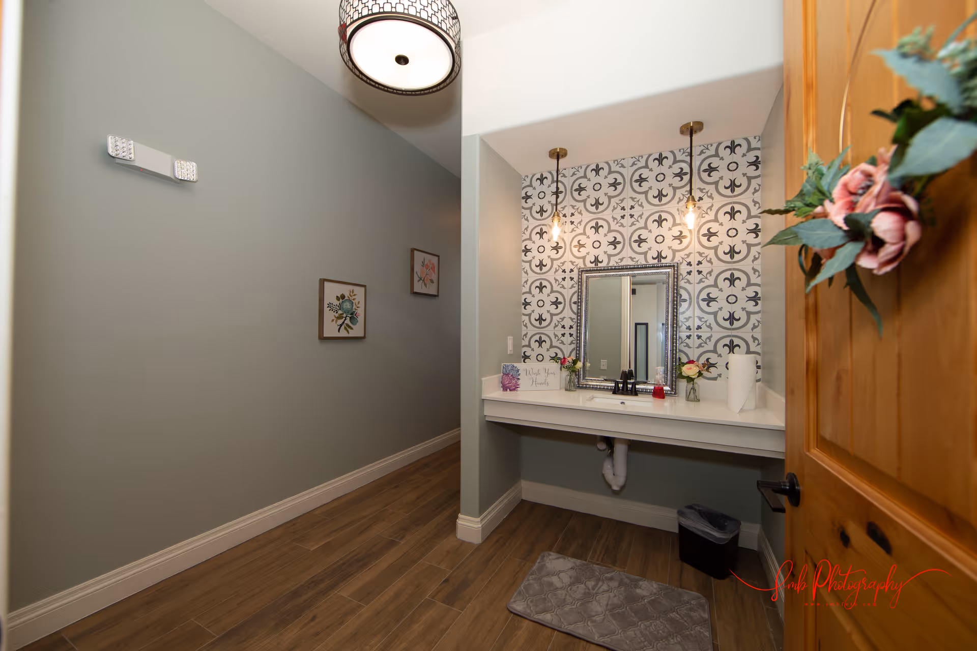 A small bathroom or wash area with a single sink, a decorative mirror, and patterned tile backsplash. The room has wooden flooring, two hanging pendant lights above the sink, and a wooden door with a floral wreath. There are two small framed floral pictures on the adjacent wall and a gray floor mat in front of the sink.