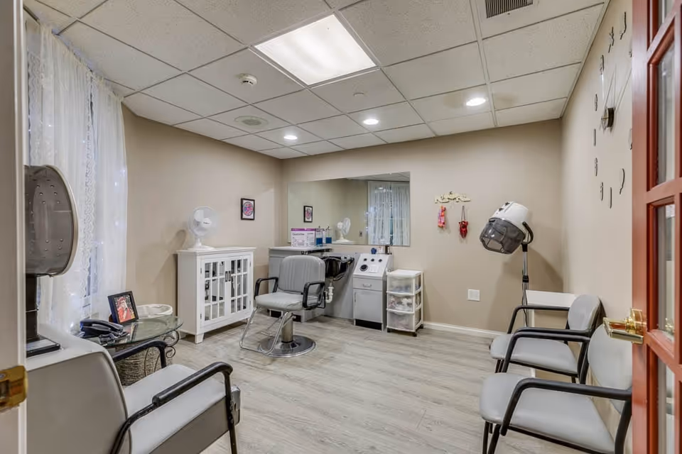 Interior view of a small salon room in an assisted living facility with beige walls and light wood flooring. The room contains salon chairs, a hair washing station, a hair dryer, a small white cabinet, a glass-top side table with a photo frame, and a large mirror on the wall. The room is softly lit with recessed ceiling lights and has a window with sheer white curtains.