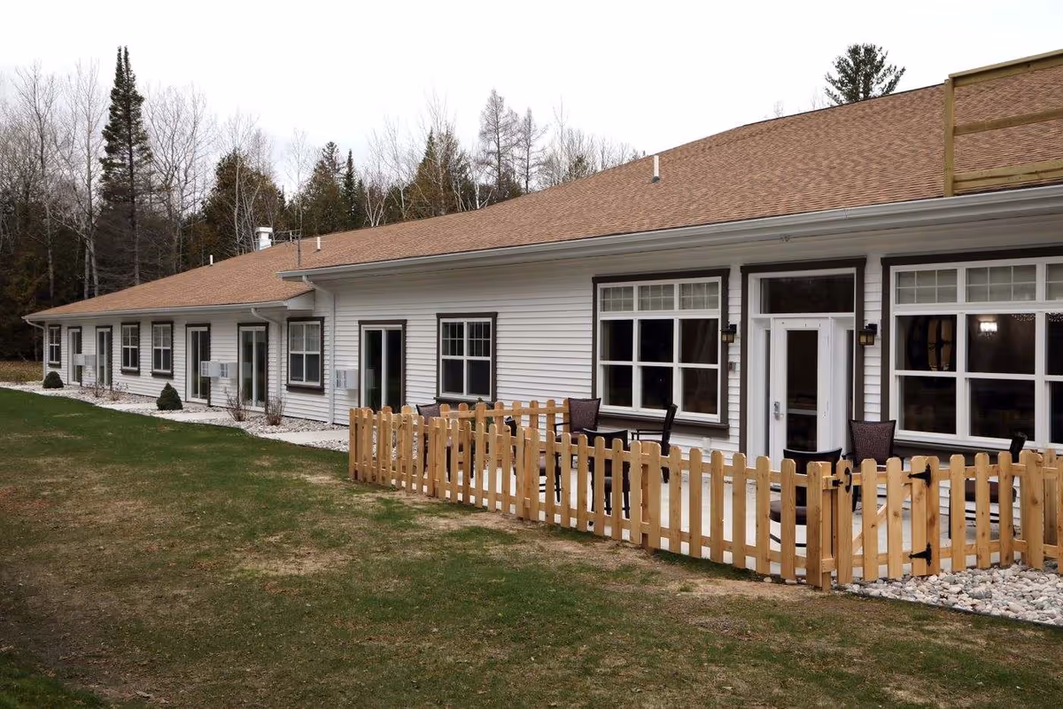 Exterior view of a single-story building with white siding and a brown roof, featuring multiple windows and doors. There is a small fenced patio area with outdoor chairs in front of the building, and trees are visible in the background.