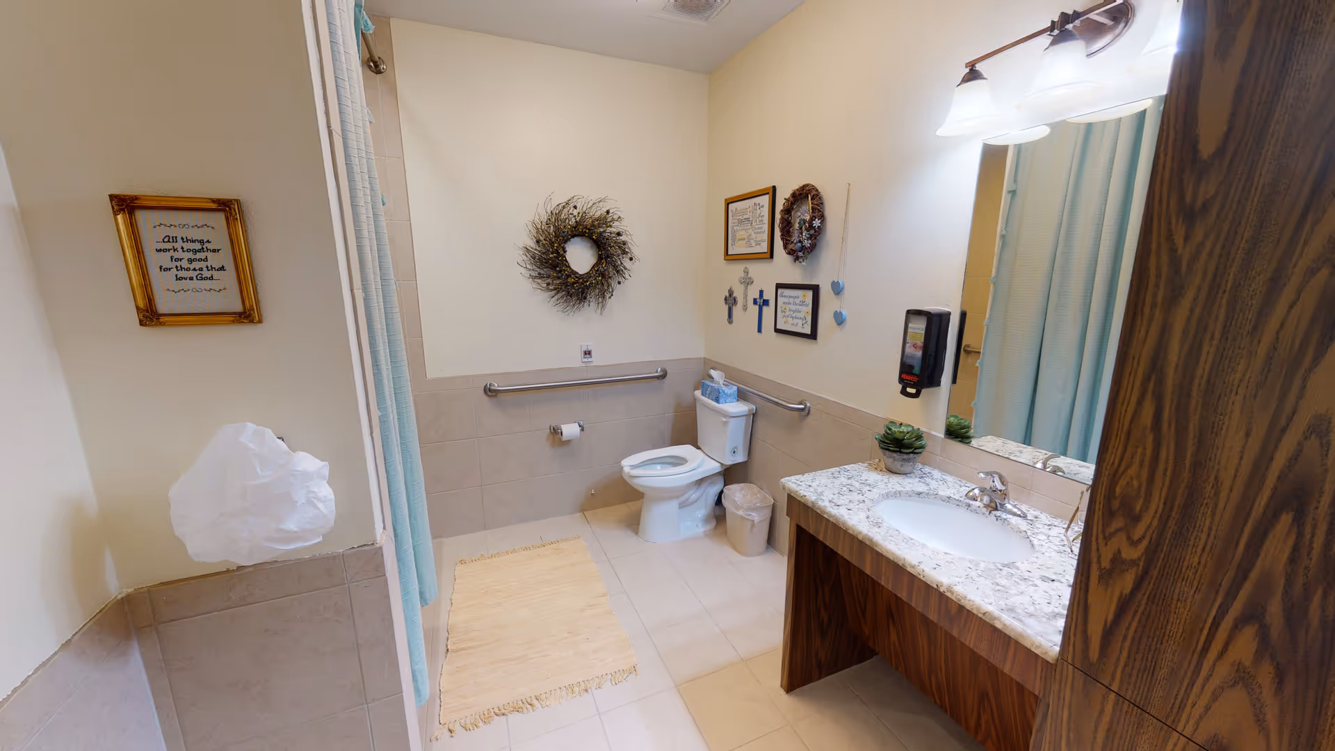 A clean and accessible bathroom with a toilet, grab bars, a sink with a granite countertop, a large mirror, and a shower with a light blue curtain. The walls are decorated with framed quotes, crosses, and a wreath. There is a beige rug on the tiled floor and a small trash can next to the toilet.
