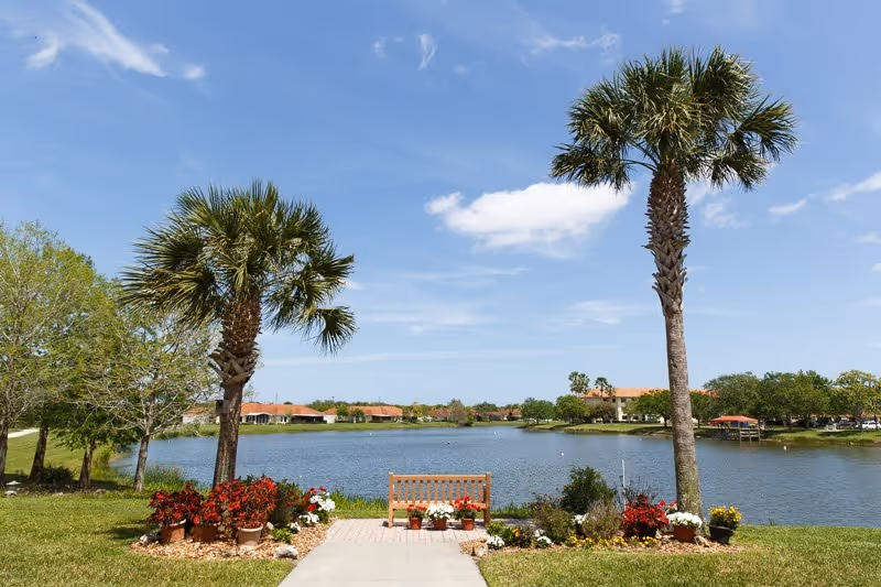 A peaceful outdoor scene at Indian River Estates featuring a wooden bench surrounded by colorful potted flowers, two tall palm trees, a calm lake, and houses with red roofs in the background under a blue sky with some clouds.