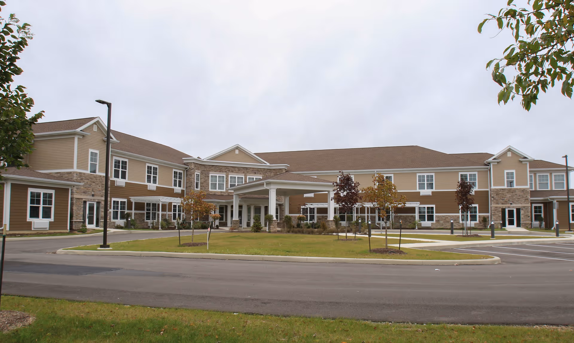 Exterior view of a two-story senior living facility building with beige and brown siding, stone accents, multiple windows, and a covered entrance. The surrounding area includes a paved driveway, small trees, and a grassy lawn under an overcast sky.