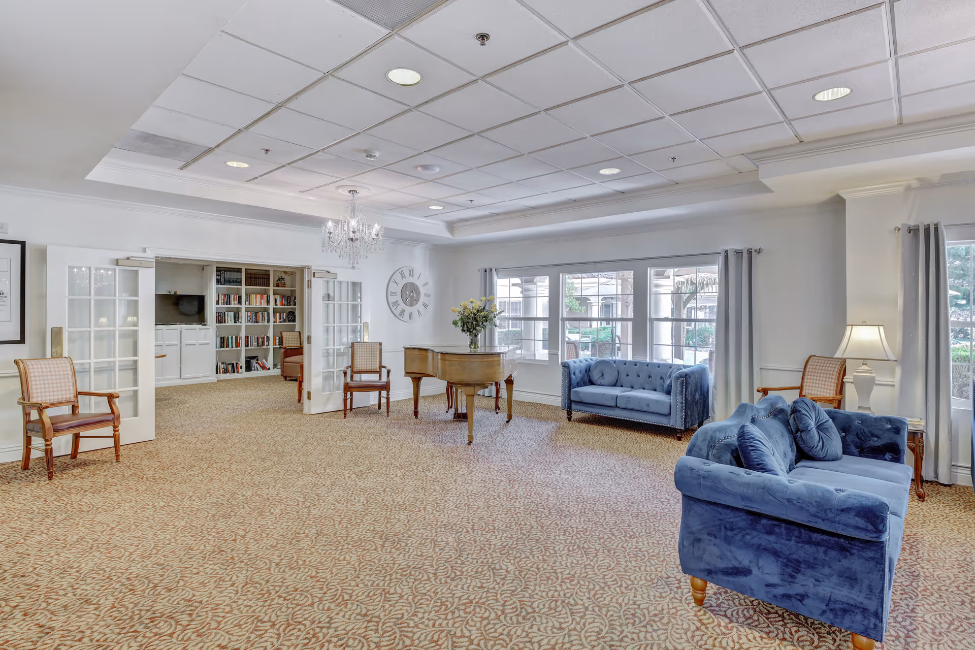 A spacious, well-lit living room area with patterned carpet, two blue velvet sofas, wooden chairs, a grand piano with a vase of flowers on top, large windows with curtains, a chandelier, and a wall clock. French doors open to an adjoining room with bookshelves and additional seating.
