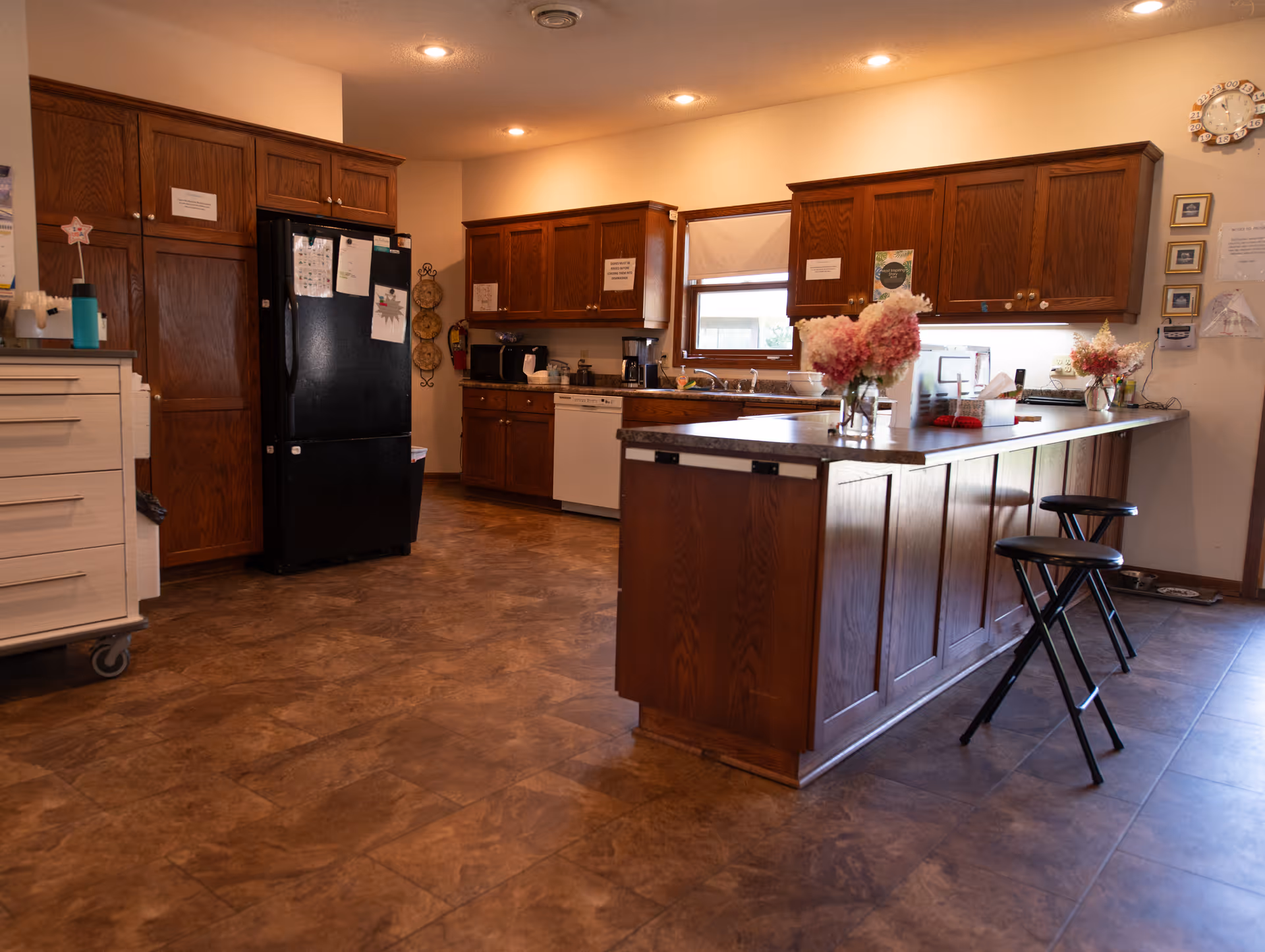 Spacious communal kitchen with wooden cabinets, a central island with stools, a black refrigerator, and floral centerpieces on the counter.