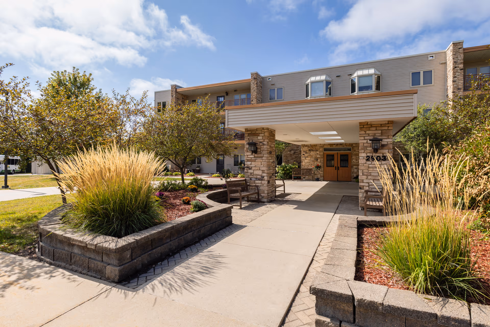 Entrance to a senior living facility with a covered drop-off area supported by stone pillars, surrounded by landscaped greenery including ornamental grasses and small trees under a partly cloudy sky.