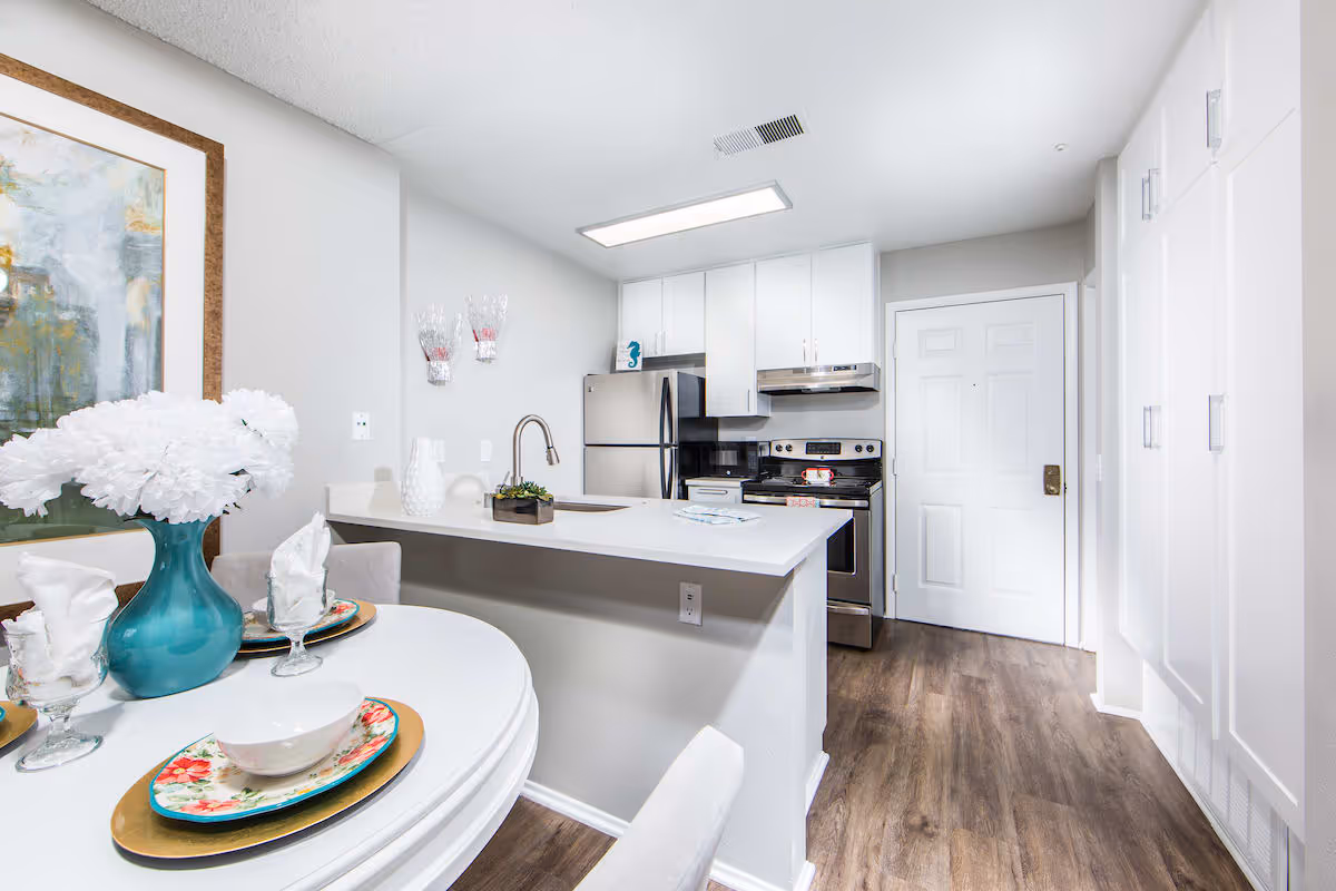 Bright and modern kitchen area with white cabinets, stainless steel refrigerator and stove, a white countertop with a sink, and a small dining table set with floral plates, glassware, and a blue vase with white flowers. The floor is wood-style, and there is a white door and built-in storage cabinets along the right wall.