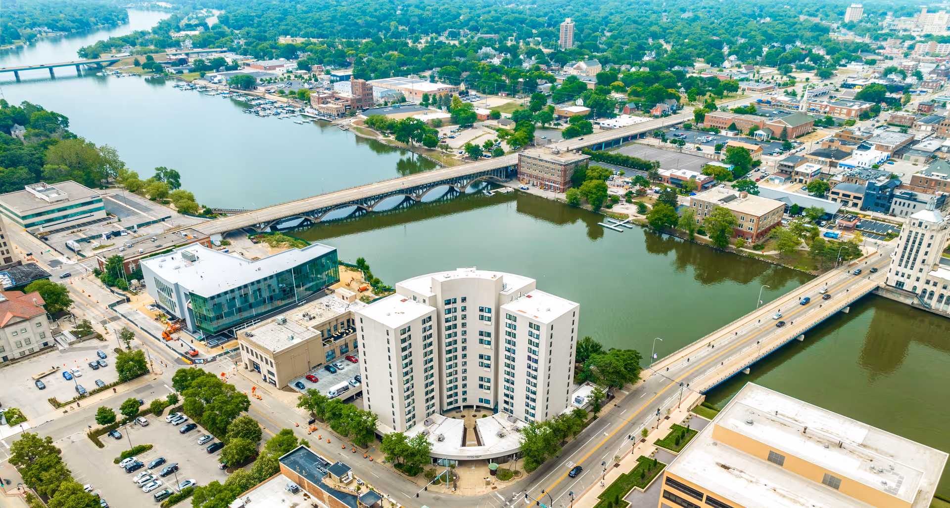 Aerial view of a curved multi-story riverfront building with bridges, streets, and surrounding city blocks.