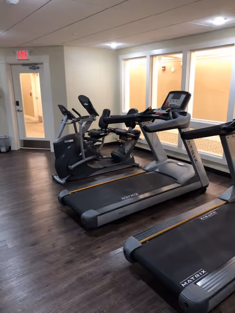 Indoor fitness room with two treadmills and two stationary exercise bikes on dark wood flooring. The room has white walls, a door with an exit sign above it, and large windows looking into another room.