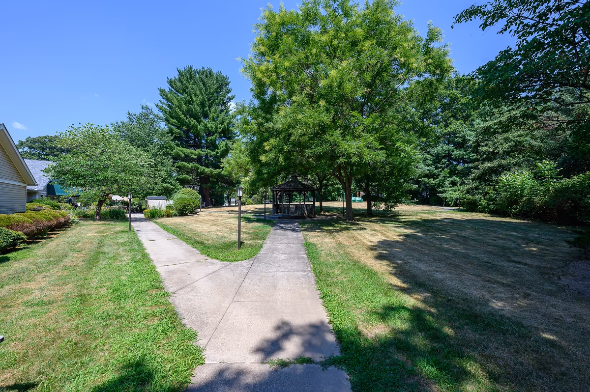 Outdoor view of a garden area with a concrete pathway splitting into two directions leading to a gazebo surrounded by green trees and grass under a clear blue sky.