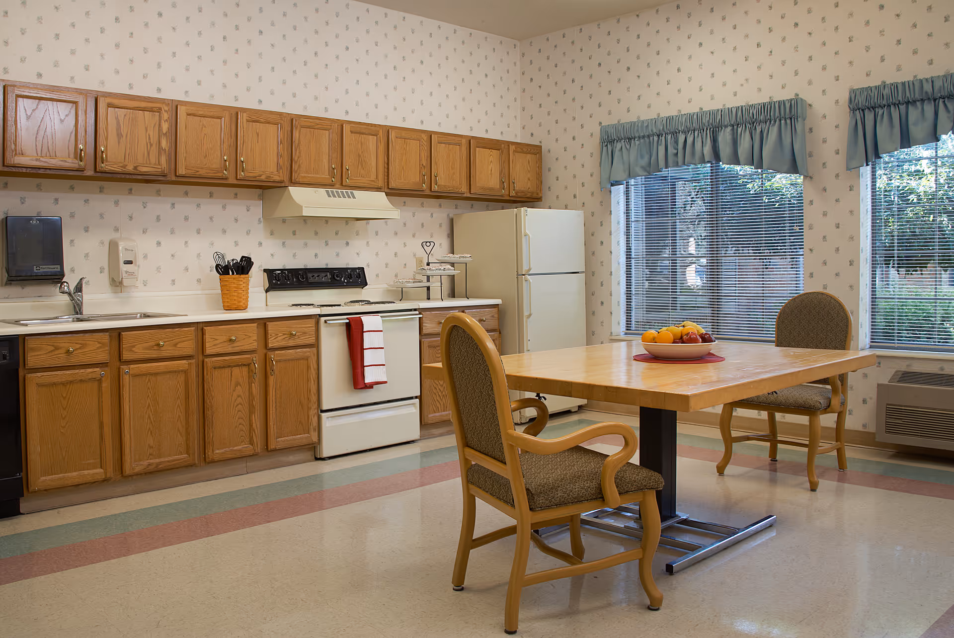 A bright communal kitchen and dining area with wooden cabinets, a stove and refrigerator, and a table with chairs and a bowl of fruit.