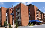 Brick multi-story retirement community building with flagpoles and a blue entrance awning.