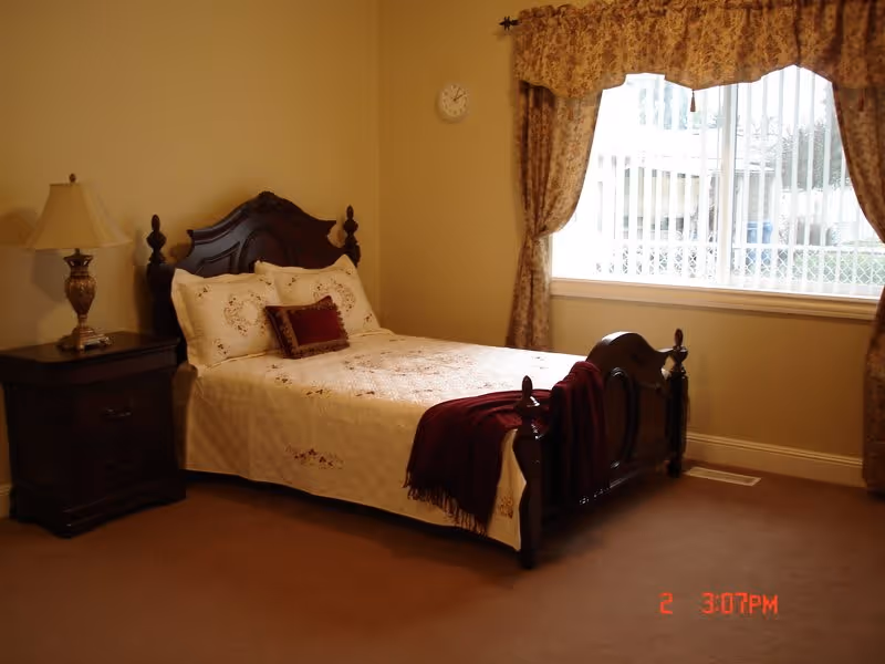 A cozy bedroom with a wooden bed frame featuring a white bedspread and decorative pillows. A burgundy throw blanket is draped over the foot of the bed. Next to the bed is a dark wooden nightstand with a classic lamp. The room has beige walls, a large window with floral curtains, and a wall clock above the bed.