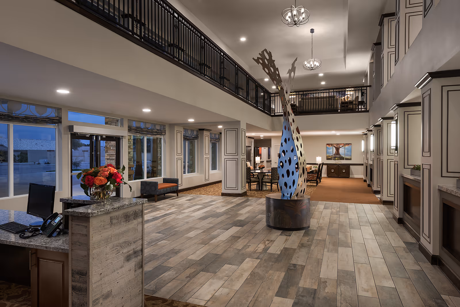 Spacious and well-lit senior living facility lobby with a modern sculpture in the center, a reception desk with a computer and phone on the left, seating areas with chairs and tables, large windows, and a second-floor balcony overlooking the space.