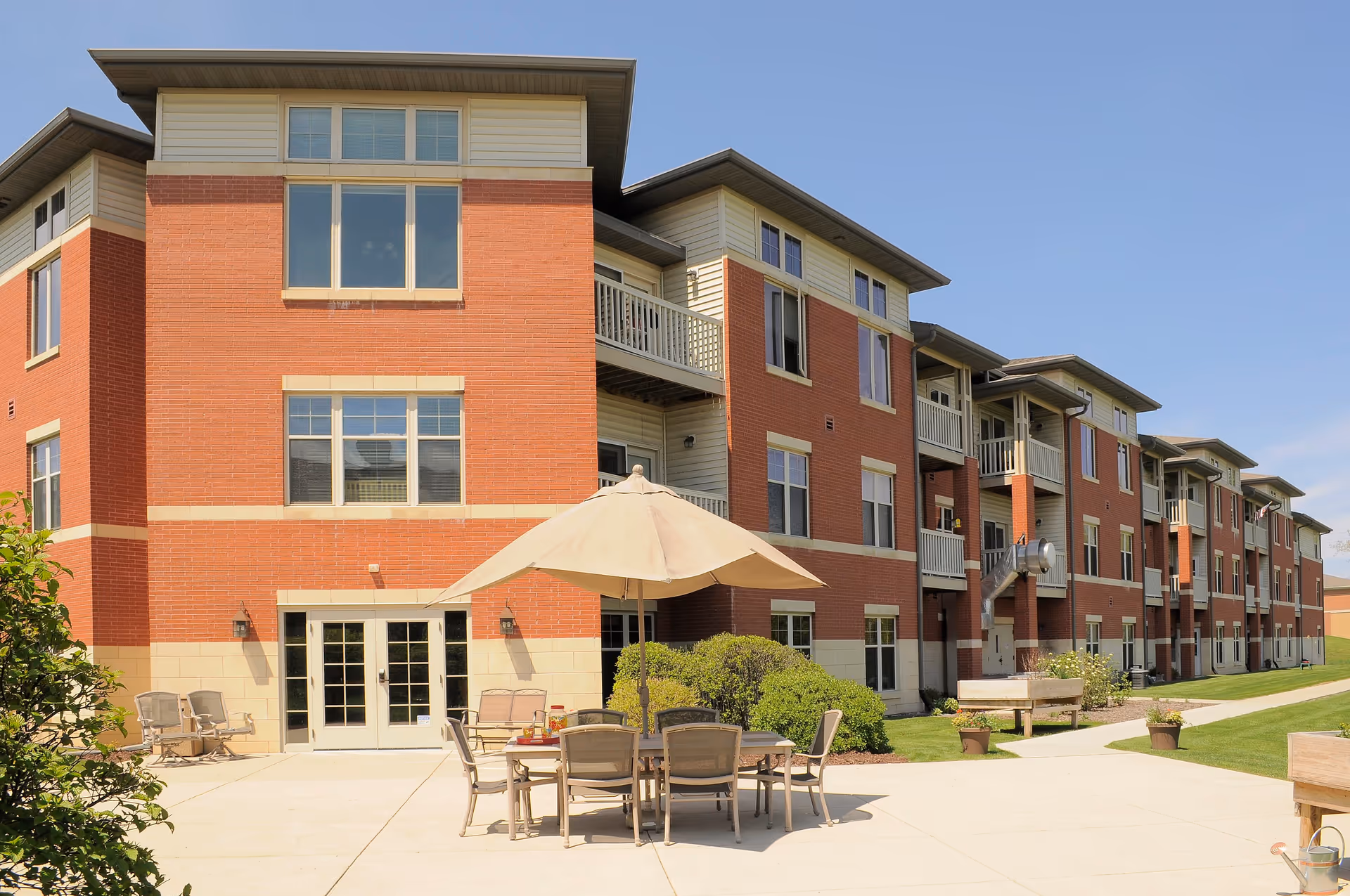 Exterior view of a multi-story senior living facility building with red brick and beige siding. There is a patio area with a table, chairs, and a large umbrella in the foreground, surrounded by greenery and a clear blue sky.