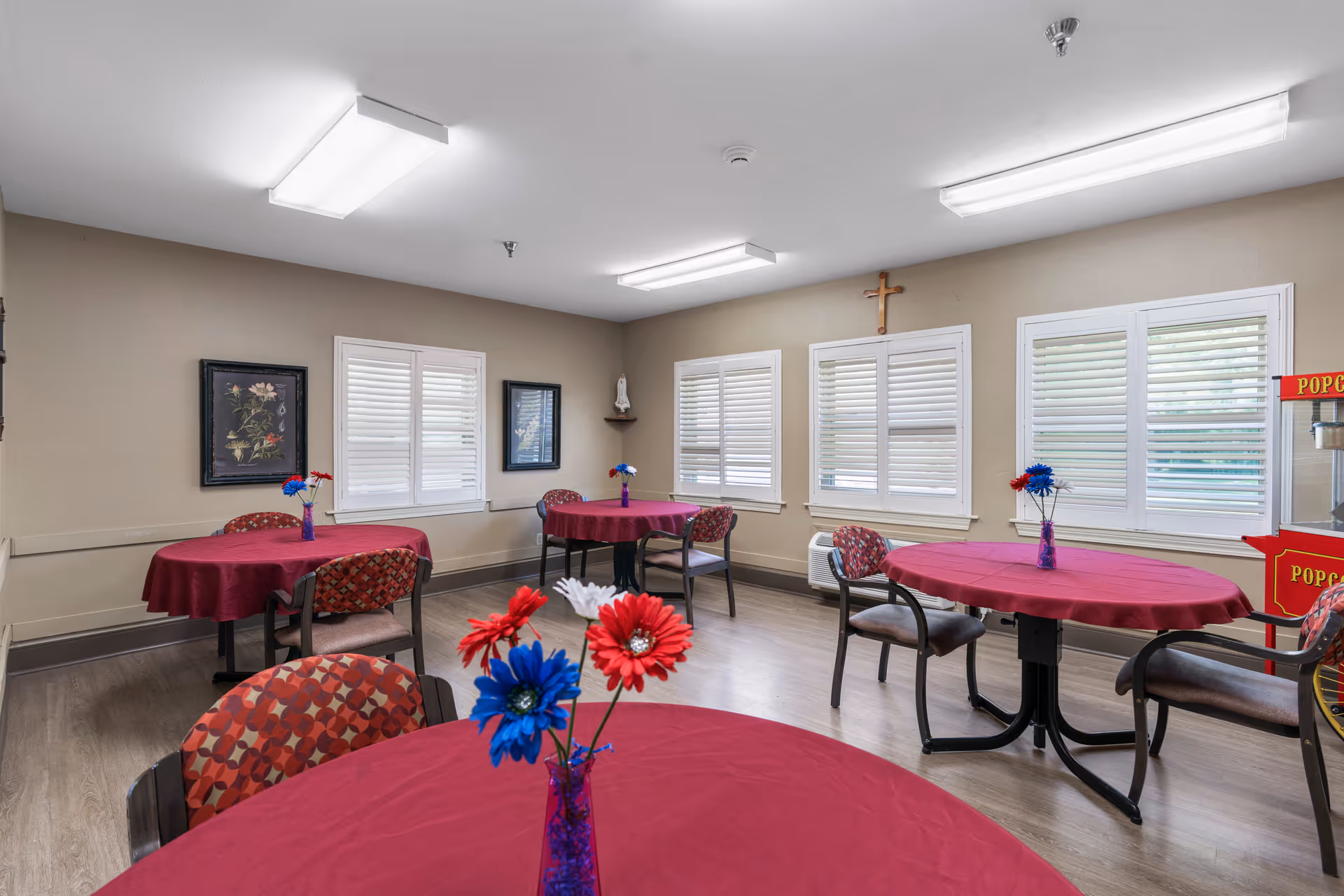 A dining room with round tables covered in red tablecloths, each table decorated with a small vase holding red, white, and blue artificial flowers. The room has beige walls, several windows with white shutters, framed floral artwork on the walls, and a popcorn machine in the corner. The floor is wood, and the ceiling has fluorescent lighting.