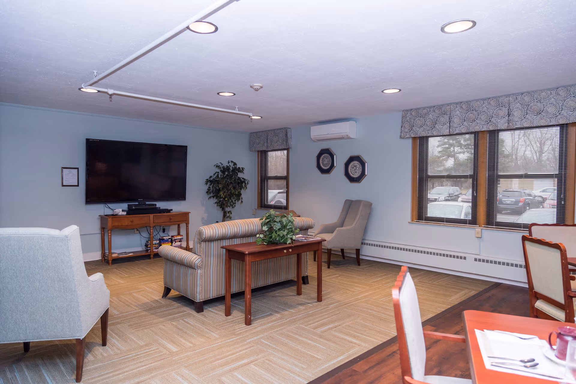 A cozy senior living community lounge area with a striped sofa, two armchairs, a wooden coffee table with a plant, a large flat-screen TV mounted on the wall, and windows with patterned valances letting in natural light. There is also a dining table with chairs partially visible in the foreground.