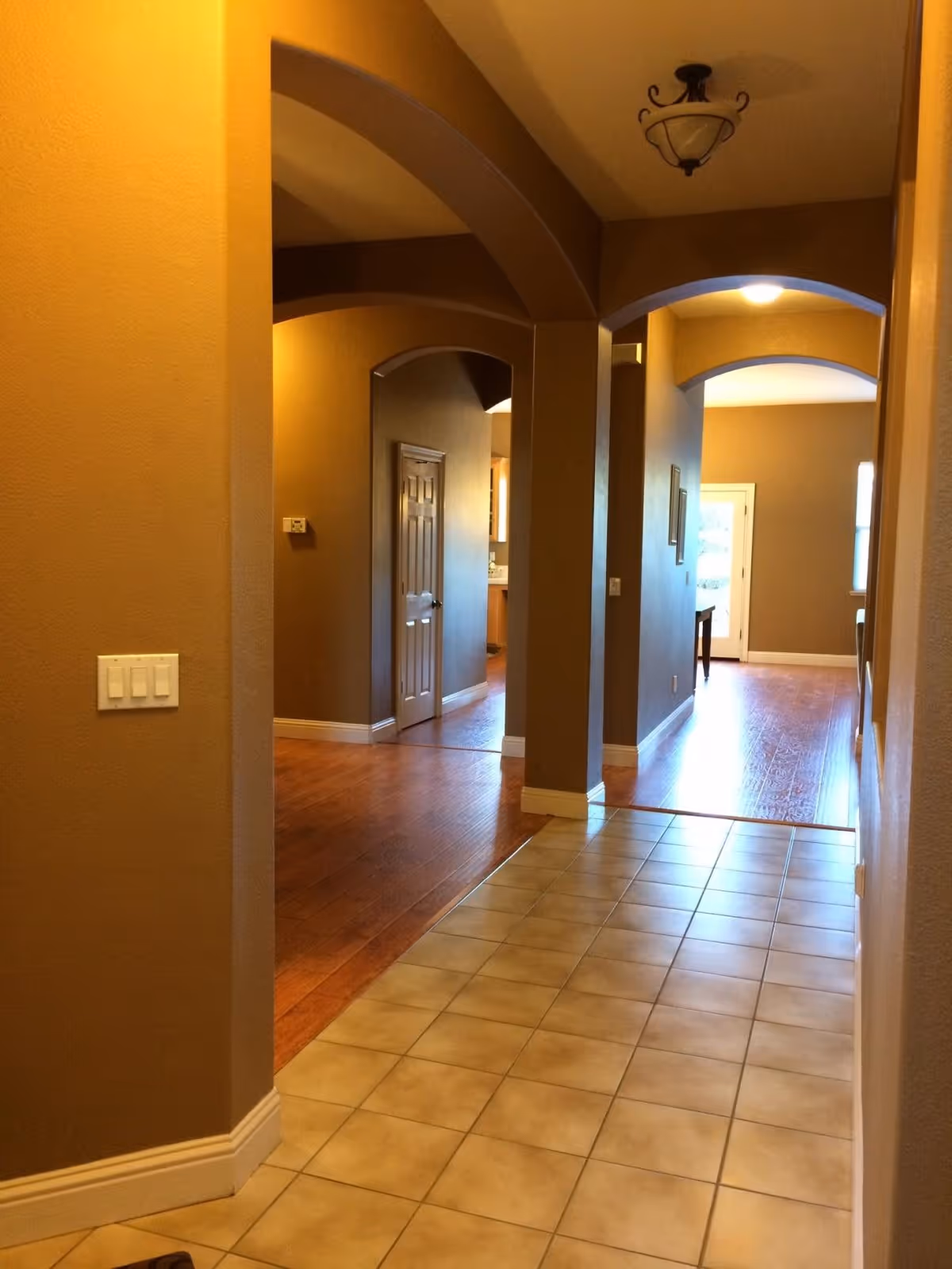 Interior hallway of a facility with beige walls and tiled floor leading to a wooden floor area. The hallway features arched doorways and ceiling light fixtures. A door and some furniture are visible in the background.