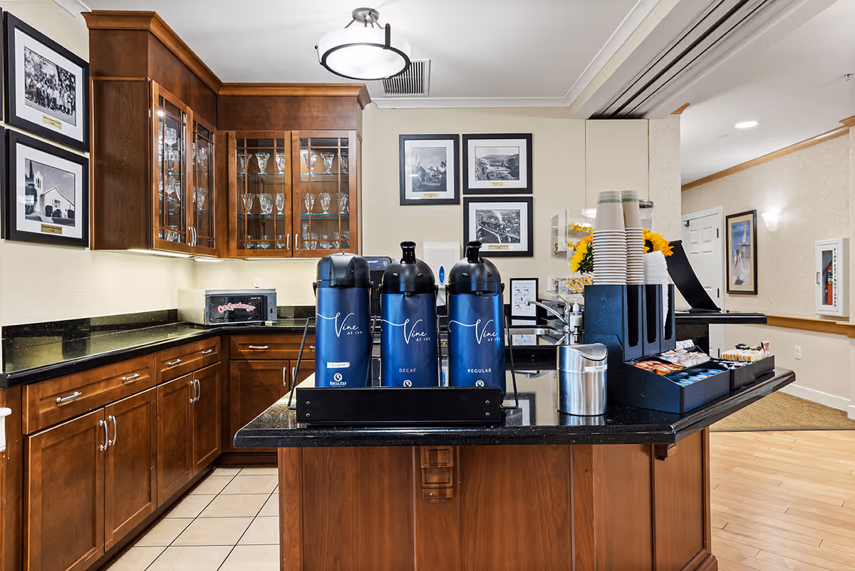 Interior view of a coffee station area in a senior living facility with wooden cabinets, black granite countertops, and three blue coffee dispensers labeled Vine at Ivy. There are stacks of disposable cups, a variety of coffee condiments, and framed black and white photos on the walls.