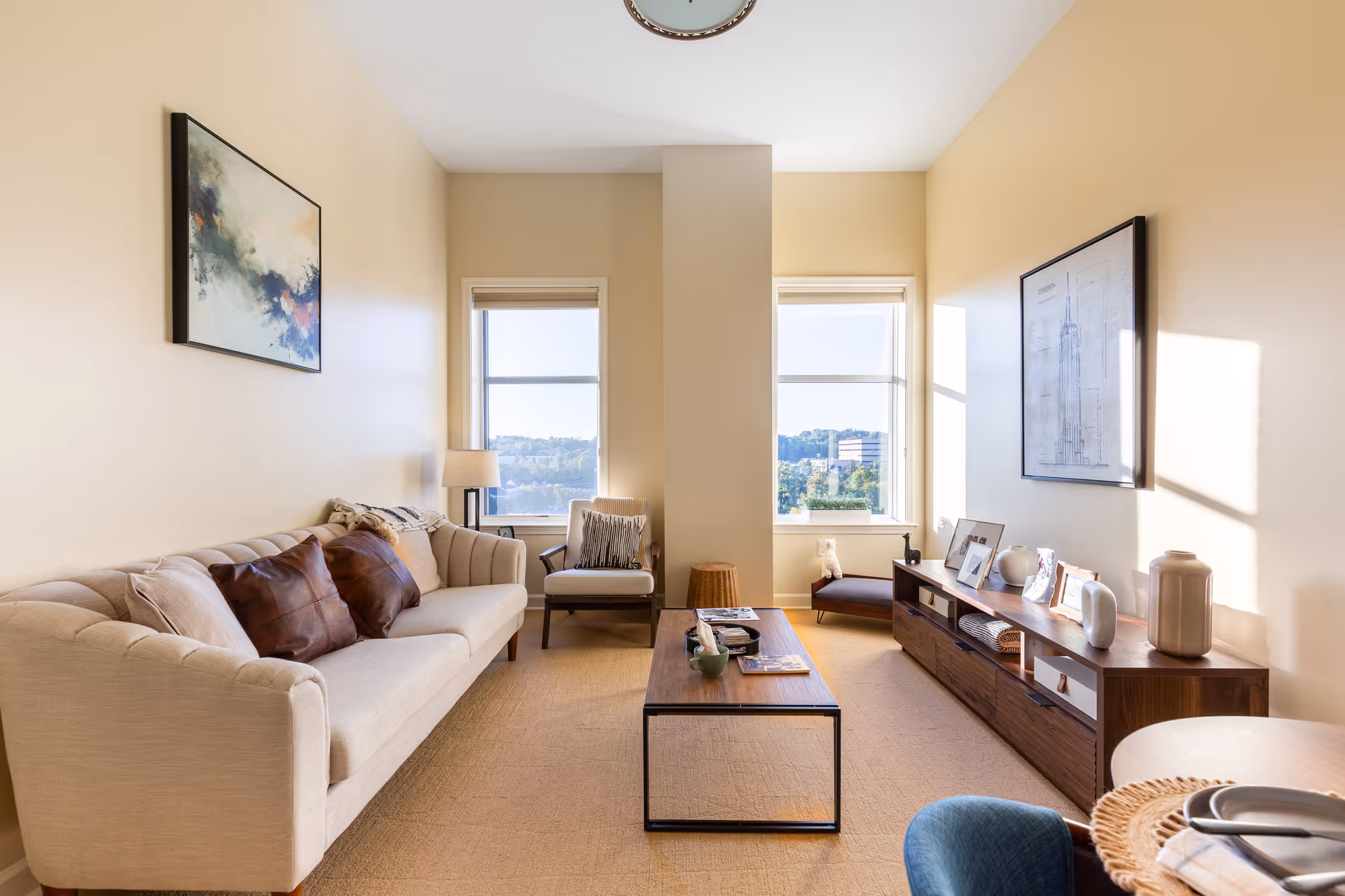 A bright living room with beige walls and carpet, featuring a cream-colored sofa with brown and beige pillows, a wooden coffee table, a single armchair near two large windows, and a long wooden console table with decorative items and framed pictures. A round wall clock is visible above the windows, and artwork hangs on the walls.