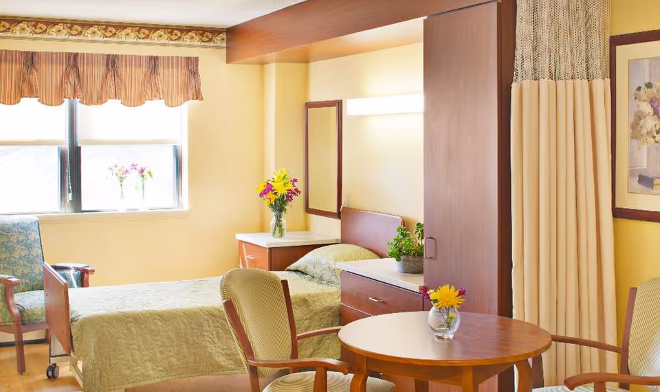 A bright and cozy bedroom in a senior living facility with a single bed covered in a light green quilt. There is a wooden nightstand with a vase of colorful flowers next to the bed, a wooden wardrobe, and a small round table with two chairs. The room has a window with striped valance curtains and a floral armchair near the window. A framed floral painting hangs on the wall, and a privacy curtain is partially visible.