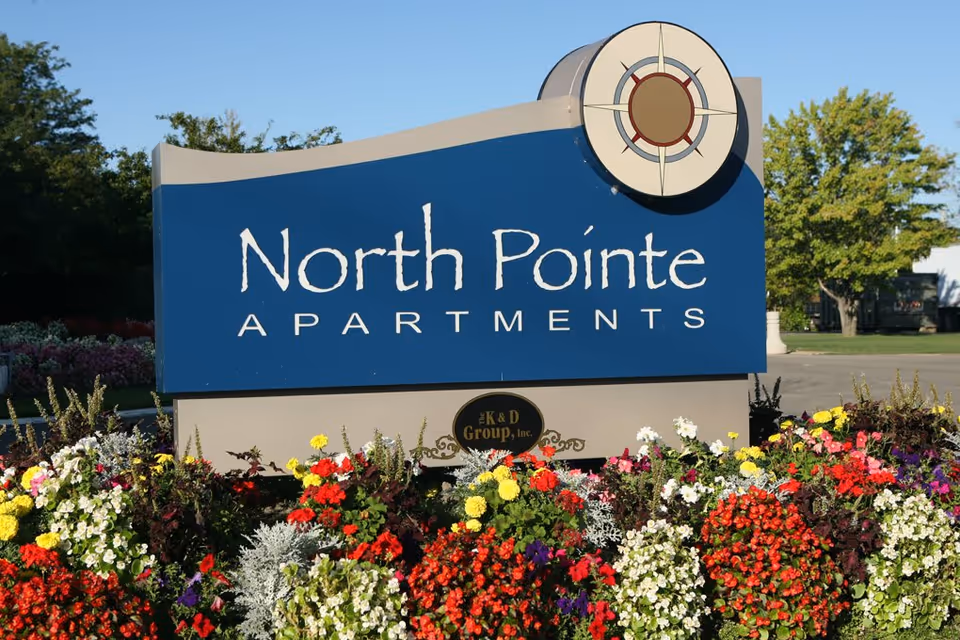 A large sign for North Pointe Apartments surrounded by colorful flowers with trees and a clear blue sky in the background.