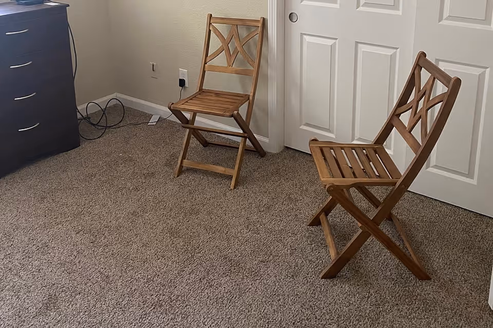 A carpeted room with two wooden folding chairs placed near a white double-door closet. A dark wooden dresser with silver handles is visible on the left side, with some cables on the floor nearby.