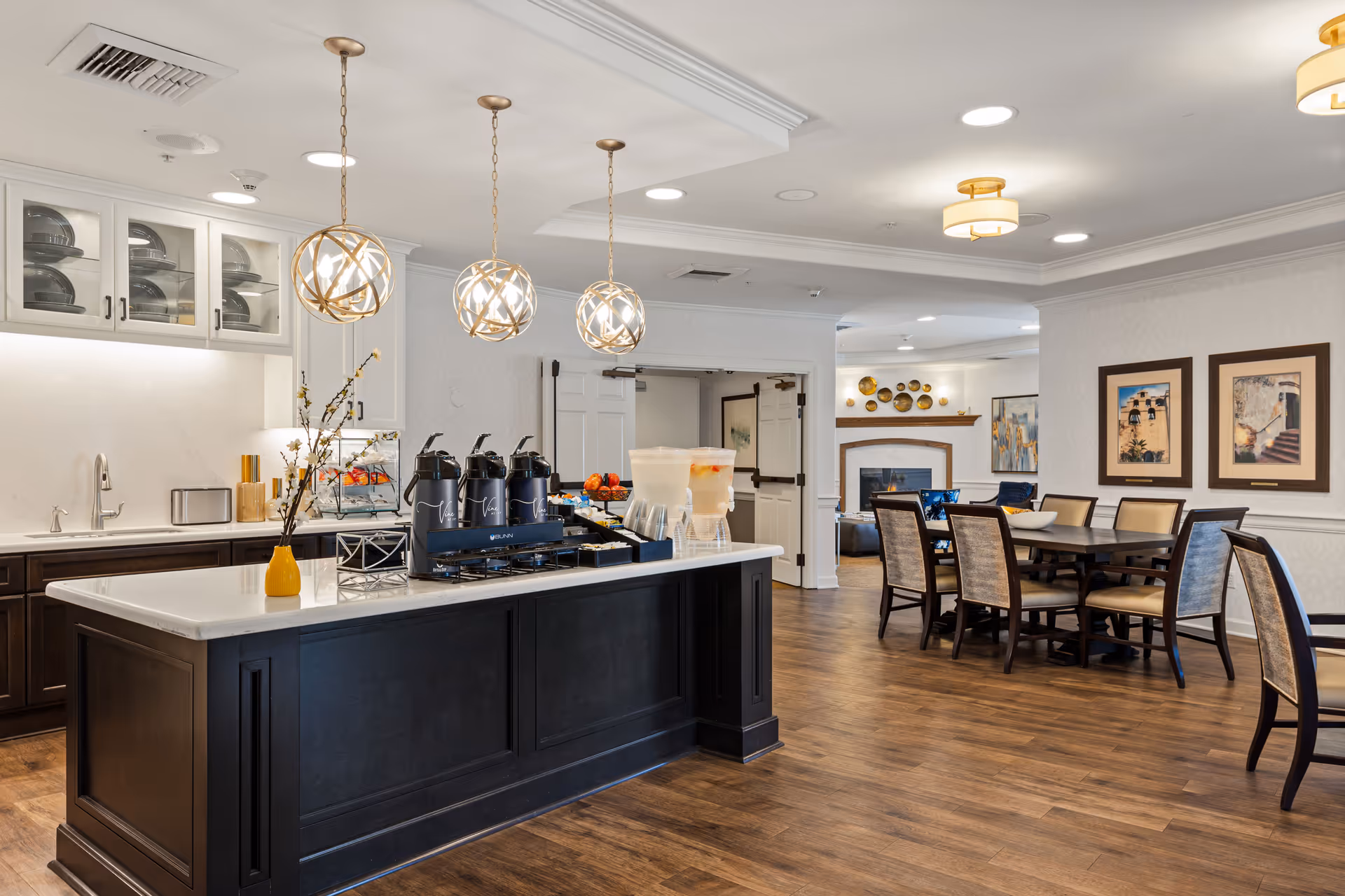 Open communal dining and kitchen area with a central island, pendant lights, beverage station, and dining table with chairs.