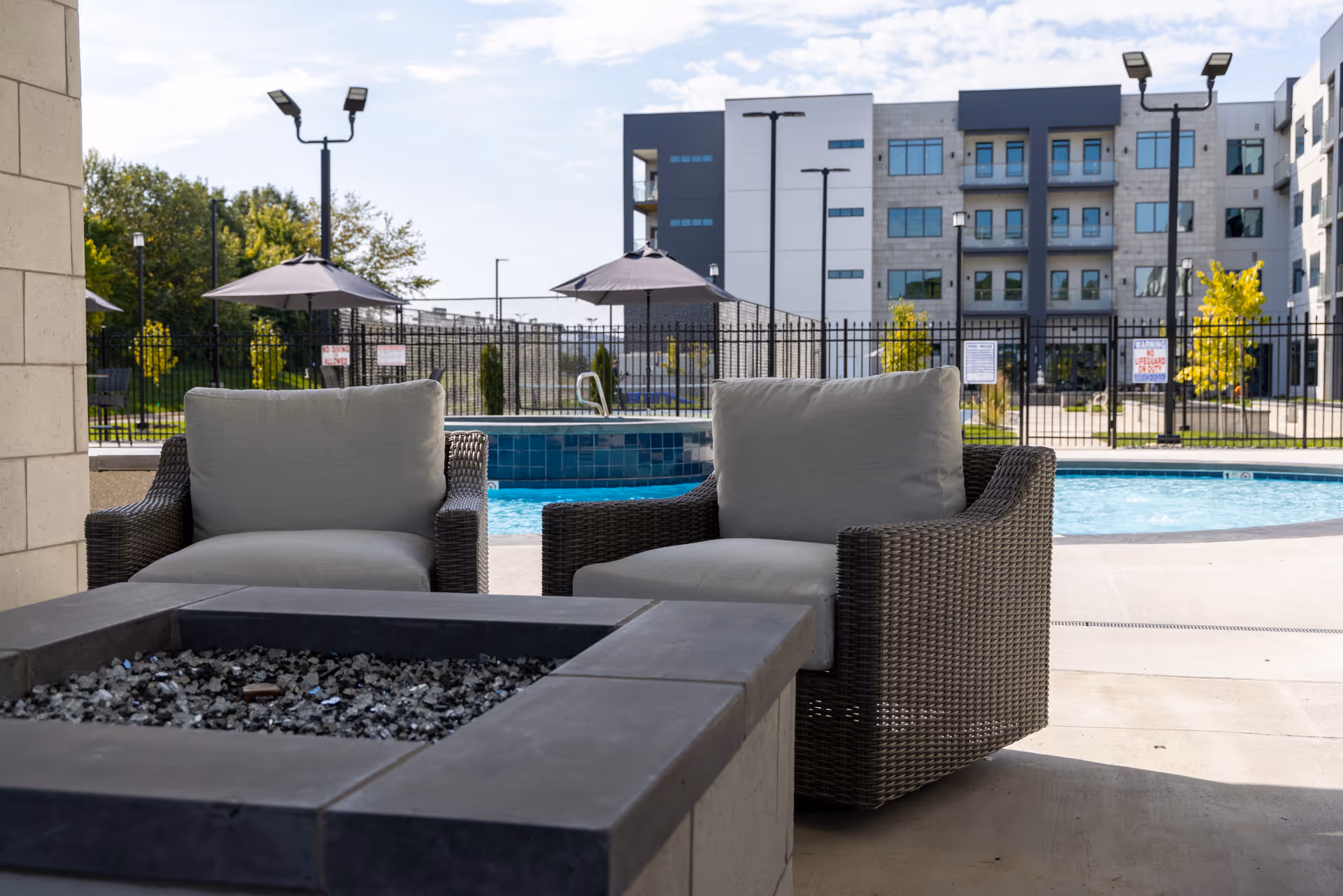 Two cushioned wicker chairs by a fire pit overlooking a swimming pool and apartment building.