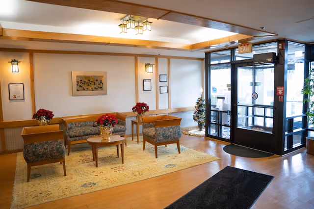 A well-lit lobby area with wooden framed floral upholstered chairs and a matching loveseat arranged around a small round wooden table with a red poinsettia plant. The floor has a large patterned yellow rug. On the right side, there is a glass entrance door with an exit sign above it and a small decorated Christmas tree nearby. The walls are light-colored with wooden trim and framed artwork and certificates.