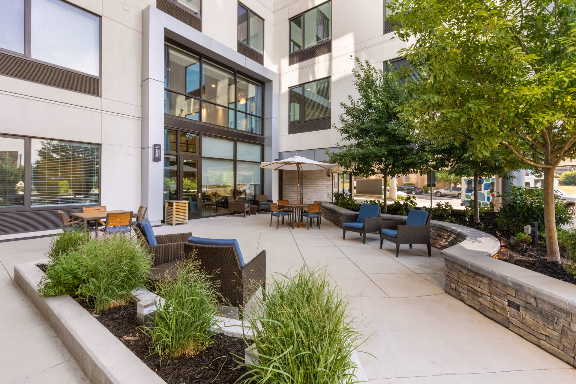 Outdoor patio area at The Belleview Senior Living with seating arrangements including chairs and tables under umbrellas, surrounded by greenery and trees, adjacent to a modern building with large windows.