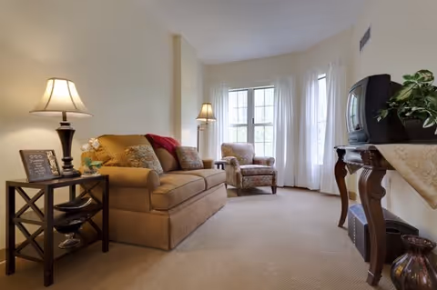 A cozy living room with a beige sofa adorned with patterned cushions and a red throw, a matching armchair near a window with white curtains, two table lamps providing warm lighting, a small side table with decorative items, and a wooden console table holding an old-style TV and a potted plant.