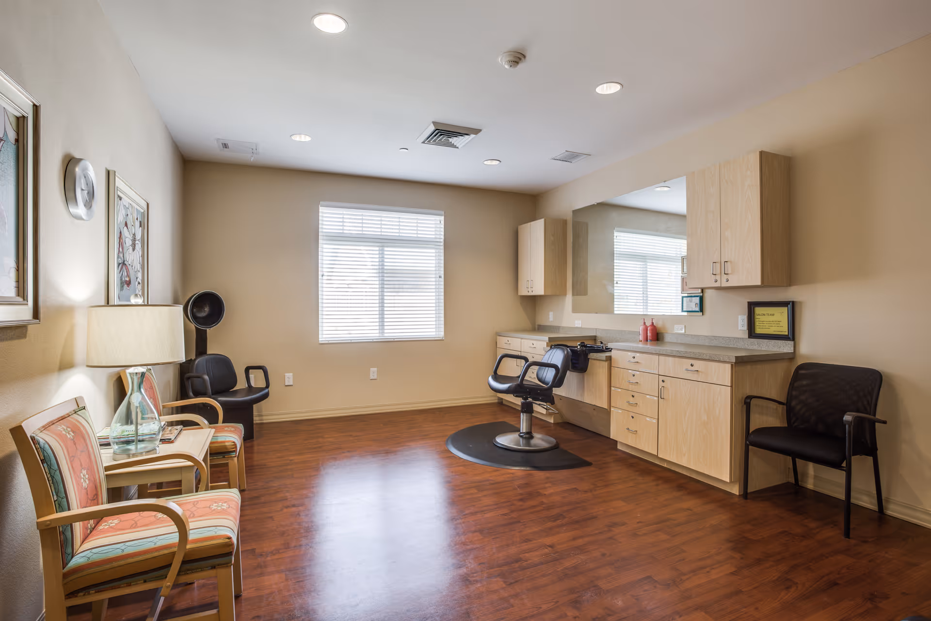 A bright room with wooden flooring and beige walls featuring a large mirror above a countertop with cabinets. There is a black salon chair on a mat in front of the mirror, a black chair to the right, and two patterned chairs with a small table and lamp to the left. A window with blinds is centered on the back wall.