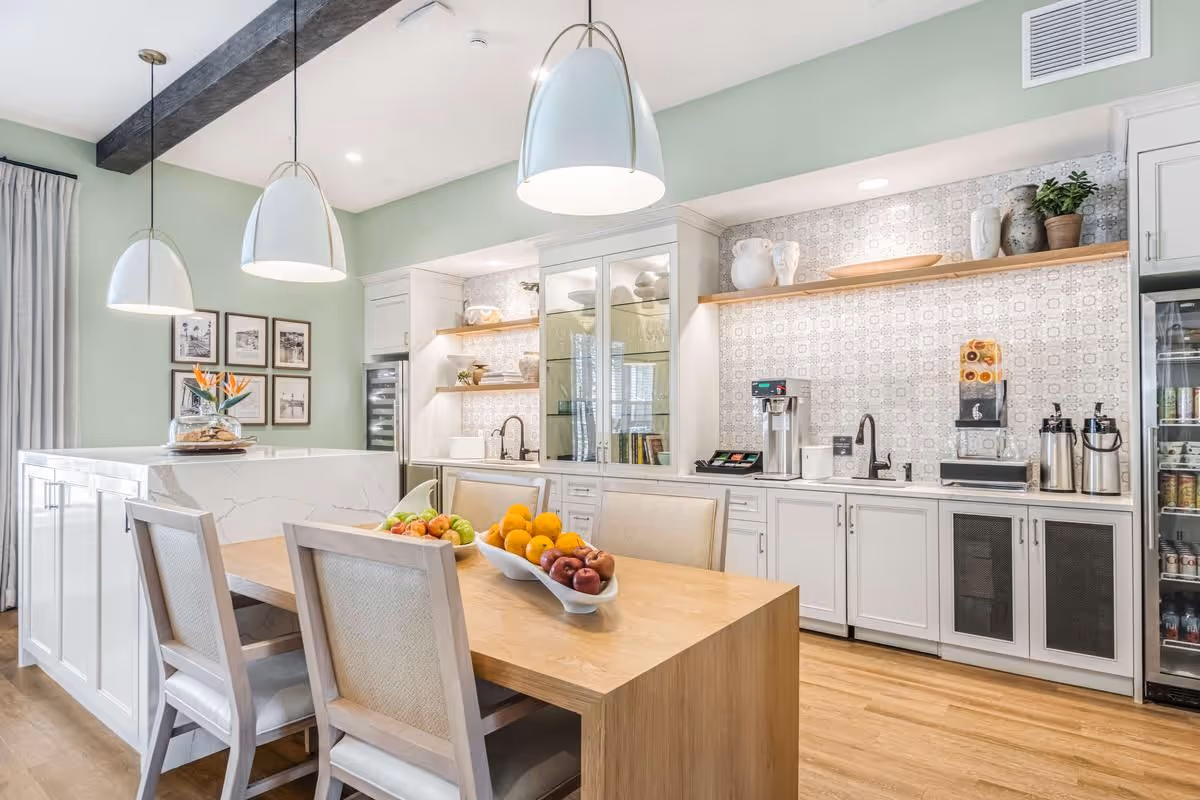 Bright communal dining and kitchenette with a wooden table topped with fruit bowls, white cabinets, pendant lights, and a beverage station.
