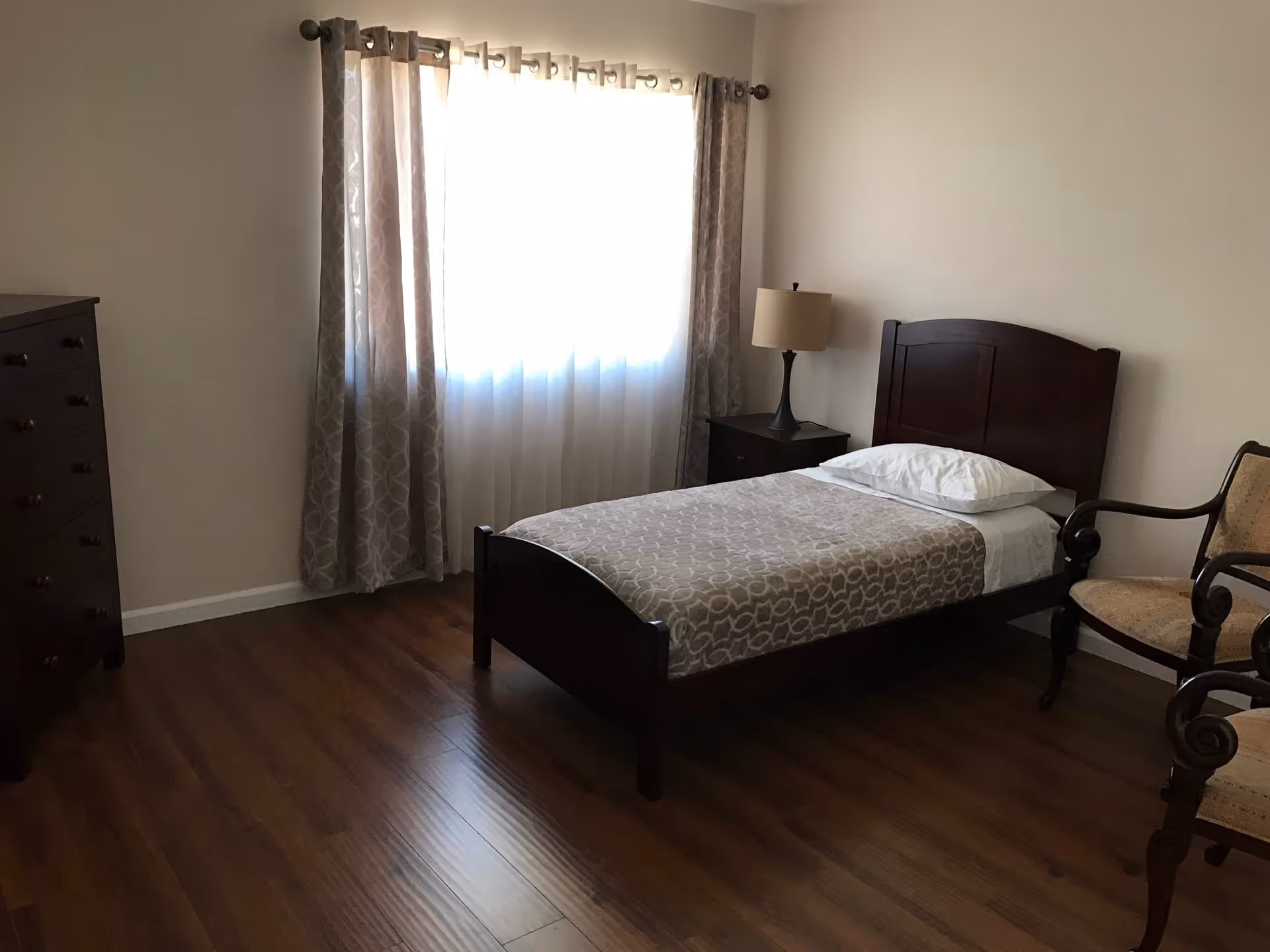A simple bedroom with a single bed covered with a patterned beige blanket and a white pillow. Next to the bed is a dark wooden nightstand with a lamp. There is a window with sheer white curtains and patterned beige drapes. A dark wooden dresser is on the left side of the room, and two wooden chairs with beige upholstery are on the right. The floor is wooden, and the walls are plain white.