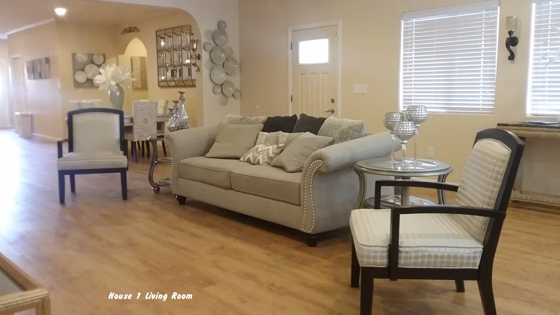 A cozy living room with a beige sofa adorned with multiple cushions, two armchairs with patterned upholstery, a small round glass side table with decorative candle holders, wooden flooring, and a window with blinds. In the background, there is a dining area with a table, chairs, and wall decorations including a mirror and plates.